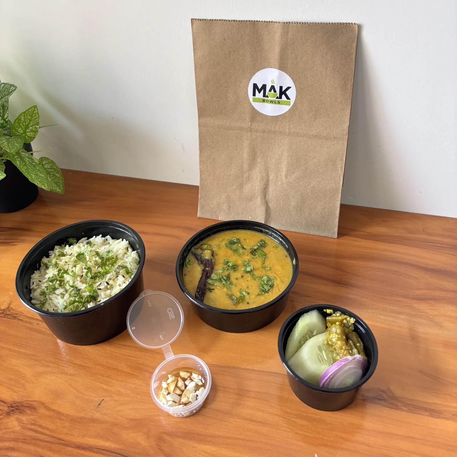 Dal tadka, jeera rice, a small side cup and a cup of salad with achar in black MAK Bowls containers, in front of a paper bag with the MAK Bowls sticker.