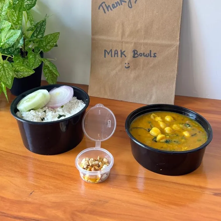 A MAK Bowls lunch on a wooden table: jeera rice topped with cucumber and onion, a yellow curry with spinach, a small side cup and a paper bag marked MAK Bowls.