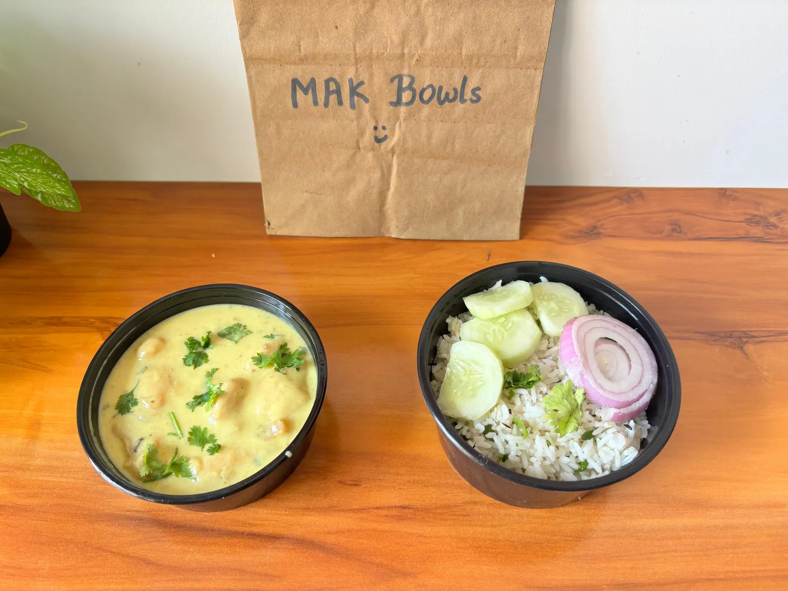 Kadhi pakora and jeera rice with cucumber and onion in two black MAK Bowls delivery containers, in front of a paper bag marked MAK Bowls.
