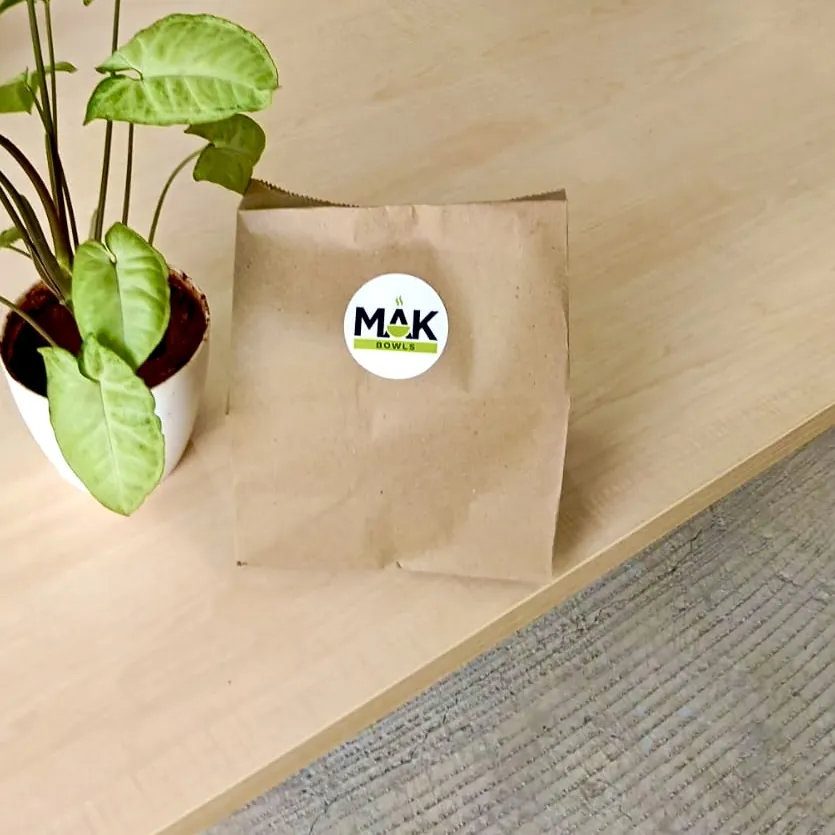 A sealed brown paper lunch bag with a MAK Bowls sticker on a wooden desk, next to a potted plant.