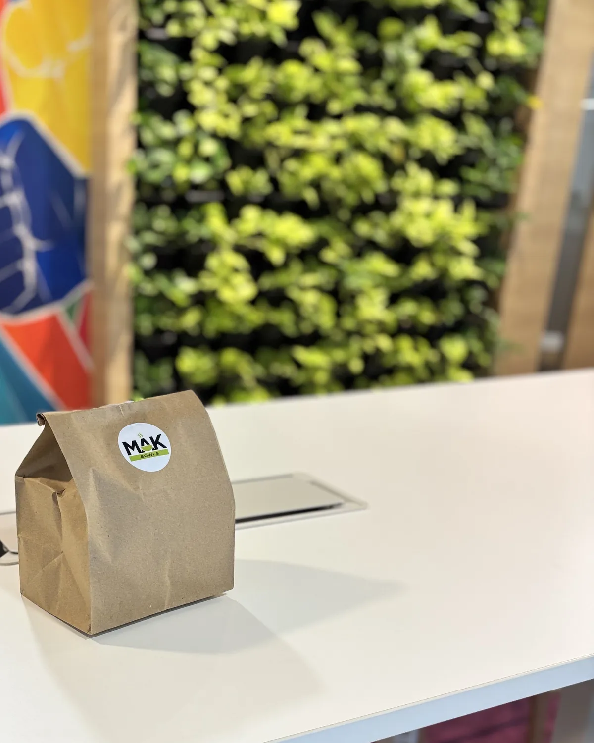 A brown paper lunch bag with the MAK Bowls sticker on a white office desk, with a green plant wall behind it.